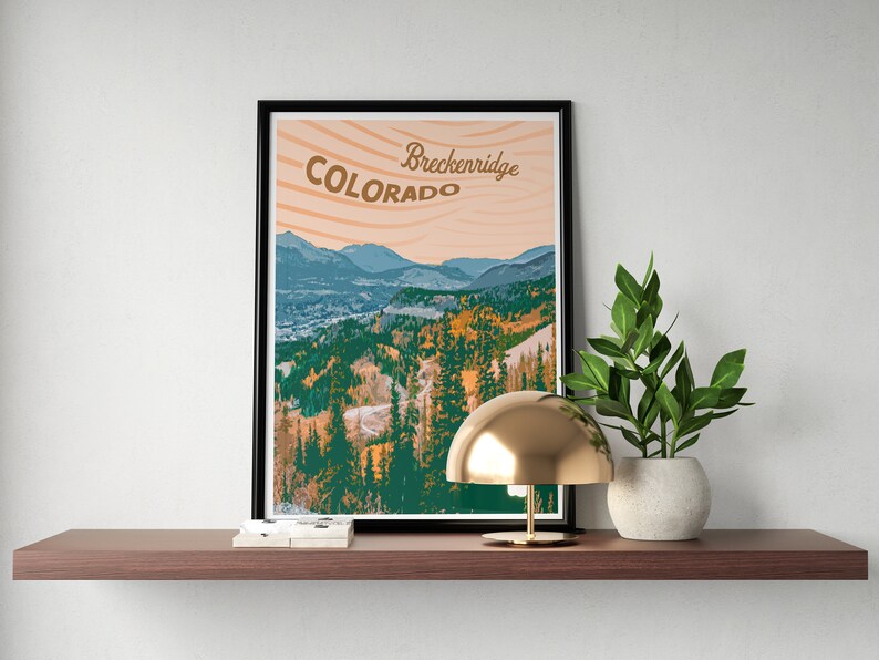 Op de afbeelding: Een ingelijste print met een berglandschap in Colorado, met de tekst "Breckenridge Colorado". De print staat op een houten plank, samen met een gouden lamp en een plant in een pot.