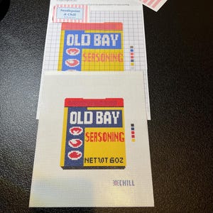 May include: Needlepoint kit featuring the Old Bay Seasoning logo. The design includes a yellow and blue box with red lettering and the text "NET WT 6 OZ". Additional materials include a note and a tag with the "Needlepoint & Chill" logo.