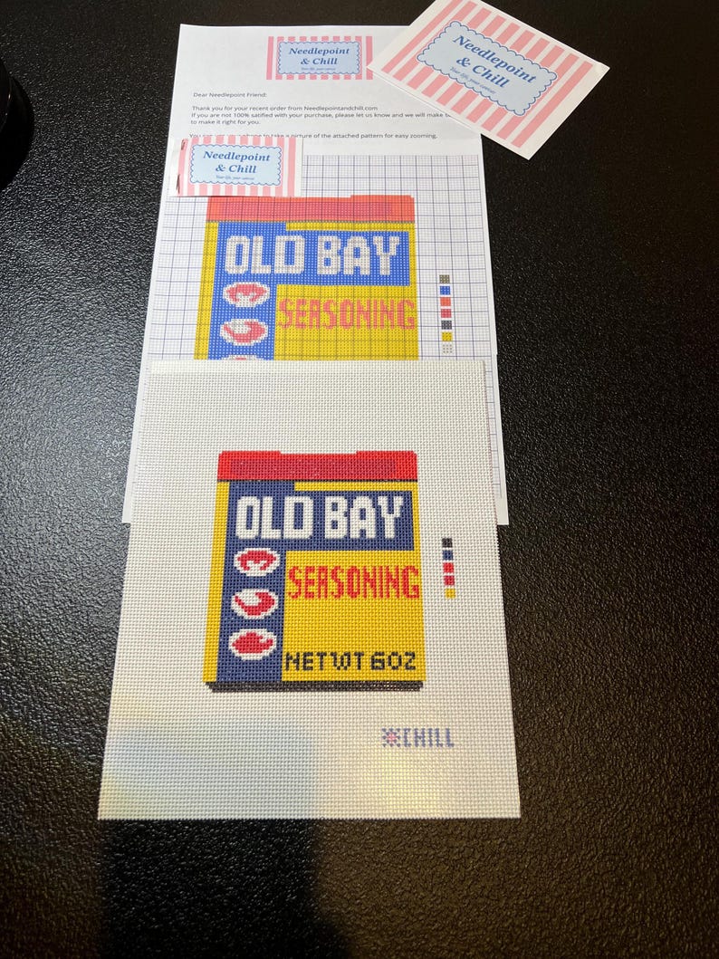 May include: Needlepoint kit featuring the Old Bay Seasoning logo. The design includes a yellow and blue box with red lettering and the text "NET WT 6 OZ". Additional materials include a note and a tag with the "Needlepoint & Chill" logo.