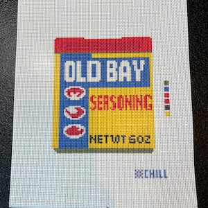 May include: A needlepoint canvas featuring a colorful depiction of an Old Bay Seasoning box. The design includes the brand name in blue and white, the word "SEASONING" in red, and the net weight of 10 oz. The word "CHILL" is also included.