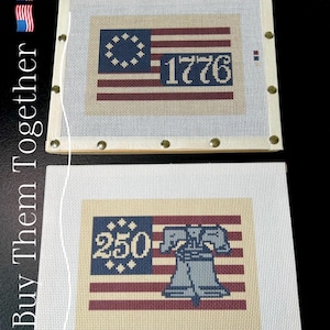 May include: Two needlepoint canvases featuring patriotic designs. One displays the American flag with "1776" and the other shows the Liberty Bell with "250". Both have a cream-colored background and are framed with wooden borders.