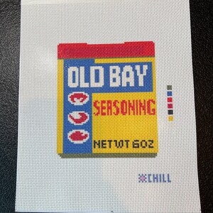 May include: A needlepoint design featuring a yellow, blue, and red box of Old Bay Seasoning. The text "OLD BAY SEASONING" is prominently displayed, with "NET WT 6 OZ" below. The word "CHILL" is also present.