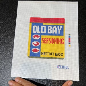 May include: A needlepoint canvas featuring a colorful depiction of an Old Bay Seasoning box. The design includes the brand name in blue and red, with a yellow background. The text "NET WT 6 OZ" is also visible, along with the word "CHILL".