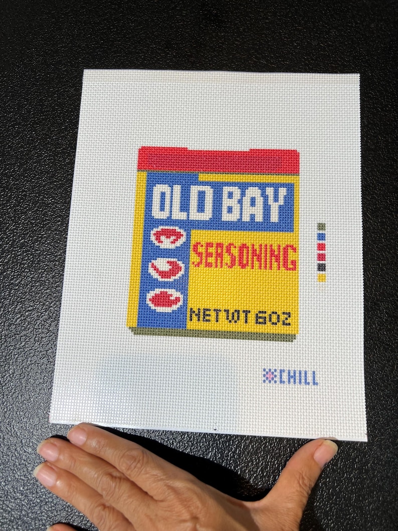 May include: A needlepoint canvas featuring a colorful depiction of an Old Bay Seasoning box. The design includes the brand name in blue and red, with a yellow background. The text "NET WT 6 OZ" is also visible, along with the word "CHILL".