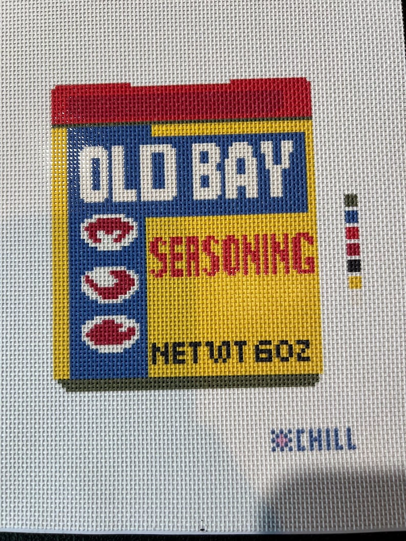 May include: Needlepoint of an Old Bay Seasoning box. The box is yellow, blue, and red with the words "OLD BAY SEASONING" in white. The net weight is 6 oz. The needlepoint is on a white background.