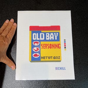 May include: Needlepoint canvas featuring a colorful depiction of an Old Bay Seasoning container. The design includes the brand name in blue and red, with the word "SEASONING" in red. The net weight is 6 oz. The background is white.