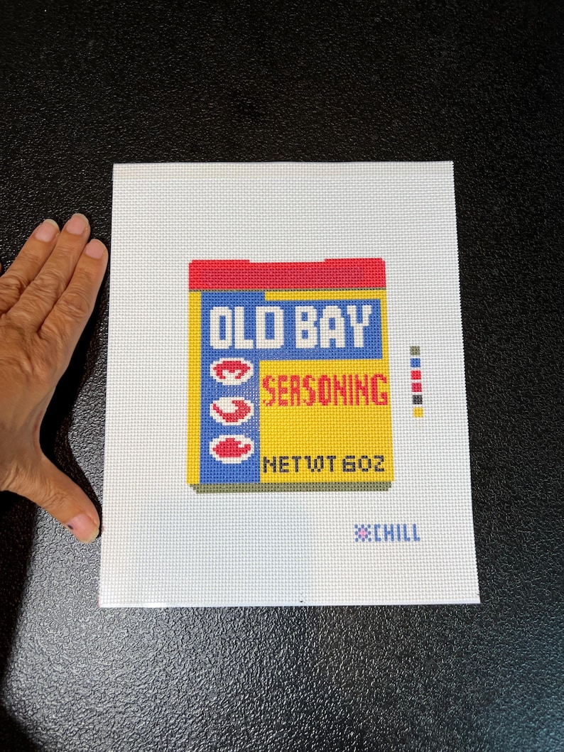 May include: Needlepoint canvas featuring a colorful depiction of an Old Bay Seasoning container. The design includes the brand name in blue and red, with the word "SEASONING" in red. The net weight is 6 oz. The background is white.