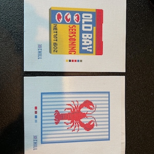 May include: Two needlepoint canvases. One features a yellow, blue, and red Old Bay seasoning box with the text "Old Bay Seasoning" and "Net Wt 6.0 oz." The other canvas shows a red lobster on a blue and white striped background.