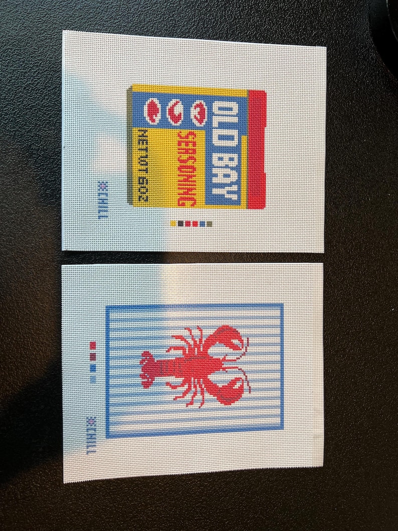 May include: Two needlepoint canvases. One features a yellow, blue, and red Old Bay seasoning box with the text "Old Bay Seasoning" and "Net Wt 6.0 oz." The other canvas shows a red lobster on a blue and white striped background.