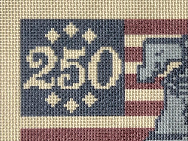 May include: Needlepoint design featuring a stylized American flag with the number "250" in white. The flag has a blue field with white stars and the number, and red and white stripes. The background is a neutral beige color.