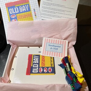 May include: A needlepoint kit featuring an "Old Bay Seasoning" design. The kit includes a canvas with the design, colored threads, a pattern, and instructions. The kit is packaged in a box with pink tissue paper and includes a note from "Needlepoint & Chill".