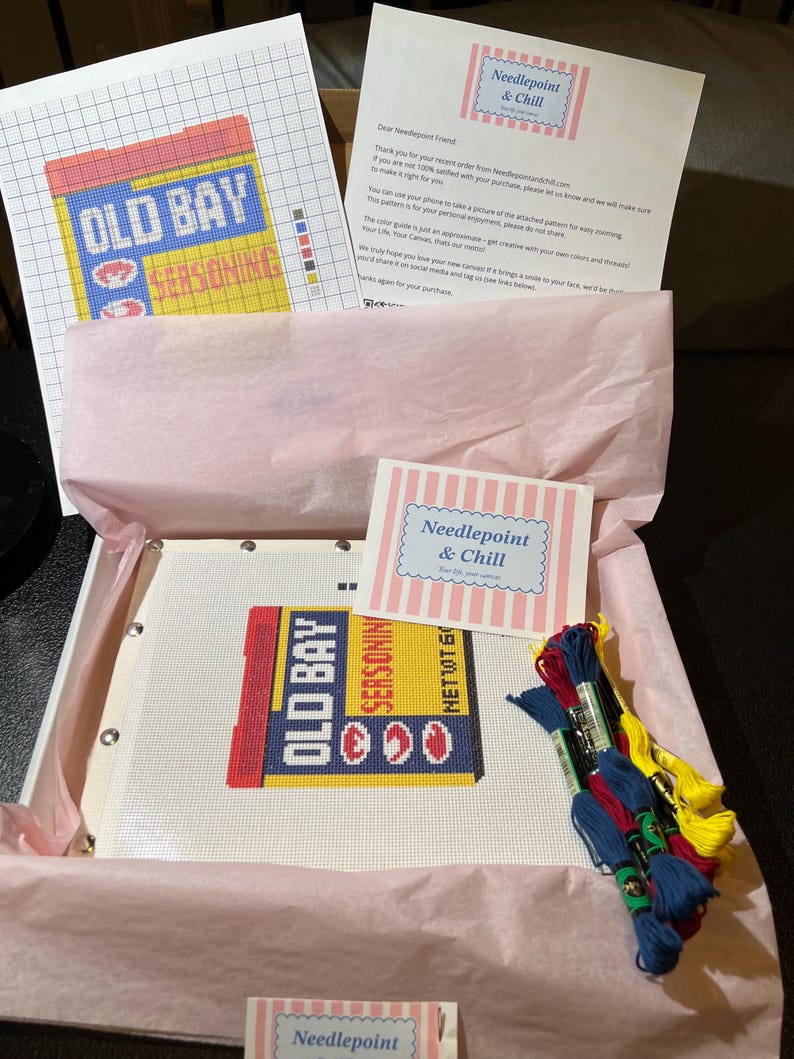 May include: A needlepoint kit featuring an "Old Bay Seasoning" design. The kit includes a canvas with the design, colored threads, a pattern, and instructions. The kit is packaged in a box with pink tissue paper and includes a note from "Needlepoint & Chill".