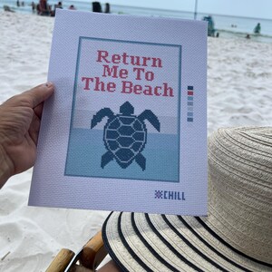 May include: A needlepoint canvas with the text "Return Me To The Beach" above a turtle design. The canvas is held on a beach with a red umbrella and a striped sun hat in the foreground.