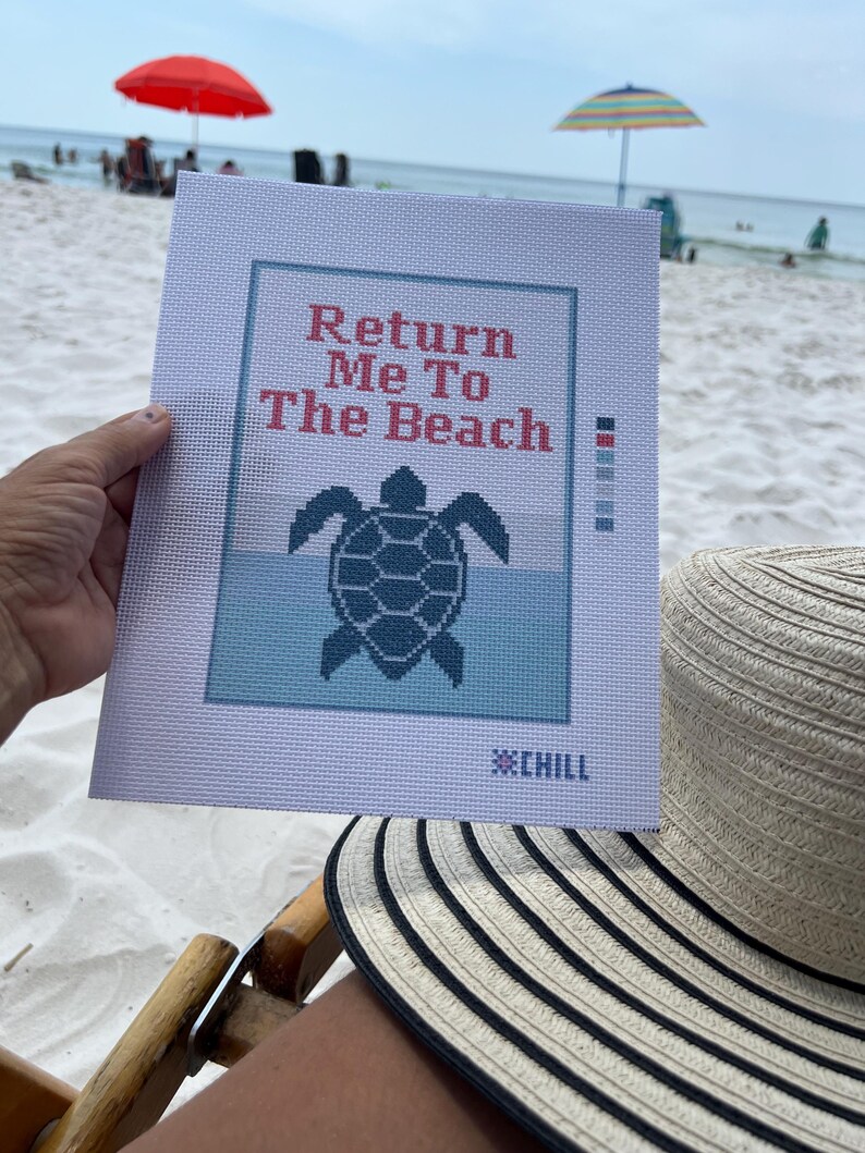 May include: A needlepoint canvas with the text "Return Me To The Beach" above a turtle design. The canvas is held on a beach with a red umbrella and a striped sun hat in the foreground.