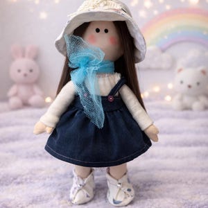 May include: A handmade doll with long brown hair, wearing a white hat, a blue scarf, and a denim dress. The doll has rosy cheeks and is wearing white shoes. A pink bunny and a white bear are in the background.