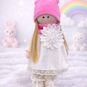 May include: A handmade doll with long blonde hair, wearing a pink hat and scarf, and a white dress with a large flower applique. The doll is standing on a white surface, with a pink bunny and a white bear in the background.