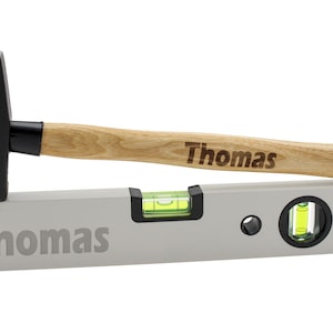 Könnte beinhalten: Ein personalisiertes Wasserwaagen- und Hammer-Set mit dem Namen "Thomas" auf beiden Werkzeugen. Die Wasserwaage ist silberfarben mit einer grünen Libelle und der Hammer hat einen Holzstiel.
