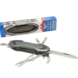 Op de afbeelding: Een zilveren multifunctioneel zakmes met een zwarte handgreep. Het mes heeft een mes, een flesopener, een schroevendraaier en andere gereedschappen. Het mes is gegraveerd met de naam "Peter". Het mes wordt getoond naast een doos met de tekst "MULTIFUNKTIONSMESSER 7 FUNKTIONEN mit 12cm Messer" erop. De doos is blauw en rood met een wit lint.
