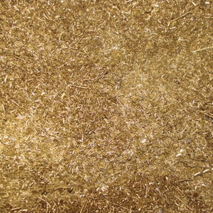 May include: Close-up of a pile of golden brass shavings. The small, irregular pieces reflect light, creating a textured, metallic surface. The image shows a dense collection of these shiny, golden fragments.