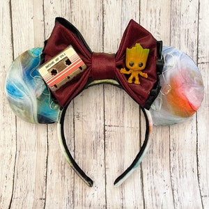 May include: A pair of mouse ears with a burgundy bow, a cassette tape, and a small Groot figure. The ears have a colorful, marbled design. The headband is black with a colorful pattern.