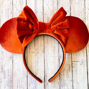 May include: Orange velvet mouse ear headband with a large bow. The ears are round and the bow is centered on the headband. The headband is black and the ears and bow are a vibrant orange.