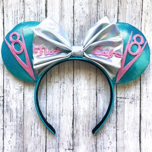 May include: A pair of mouse ears with a teal sequined design. The ears have a silver bow with the text "Flo's Cafe" in pink. The ears also feature a pink "V8" design. The headband is black.