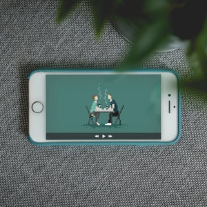 May include: A smartphone with a teal case displays a pixelated illustration of two people at a table, possibly enjoying coffee. The screen shows a play/pause bar. The phone rests on a gray textured surface, with green foliage in the background.