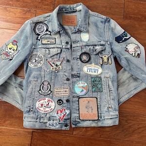 Puede incluir: Chaqueta vaquera azul claro adornada con varios parches bordados. Los parches incluyen texto, logotipos e ilustraciones. La chaqueta tiene cierre de botones, dos bolsillos en el pecho y cuello clásico. Estilo vintage.