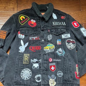 Puede incluir: Chaqueta vaquera negra con cuello de sherpa y varios parches, incluyendo Nirvana, Queen, un panda, una calavera, una guitarra y una cruz roja y blanca suiza.