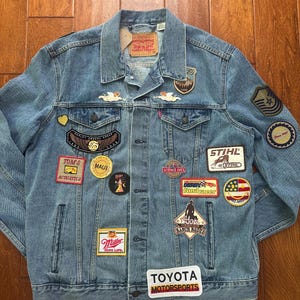 Puede incluir: Chaqueta vaquera azul claro con múltiples parches. Los parches incluyen el texto "TOYOTA MOTORSPORTS", "STIHL", "Miller" y "MAUI". Otros parches presentan varios logotipos y diseños. La chaqueta tiene un diseño clásico con botones y bolsillos.
