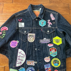 Puede incluir: Chaqueta vaquera azul oscuro adornada con varios parches bordados. Los parches incluyen texto como "BEA NICE HUMAN", "Insert Screw Here" y "LOVE", junto con imágenes de un flamenco, una abeja y un dinosaurio. La chaqueta tiene un cuello clásico y botones.