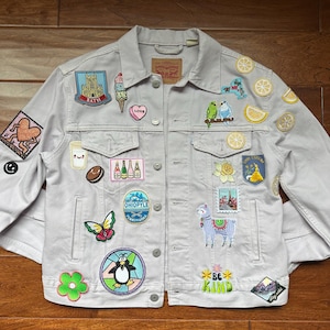 May include: A light gray denim jacket with a variety of colorful patches sewn on. The patches include a penguin, a butterfly, a llama, a flower, a lemon, a heart, a church, an ice cream cone, a bottle of wine, a donut, a bird, and more.