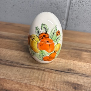 May include: A white ceramic egg-shaped salt shaker with a hand-painted design of orange and yellow fruits and green leaves. The shaker is on a wooden surface.