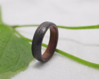 May include: A dark gray wooden ring with a brown wood inlay. The ring is sitting on a white surface with a green leaf in the background.