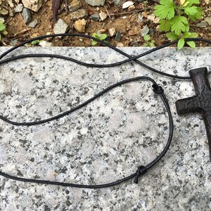 Puede incluir: Un colgante de cruz de madera negra en un collar de cord&oacute;n negro. La cruz tiene una superficie rugosa y texturizada.