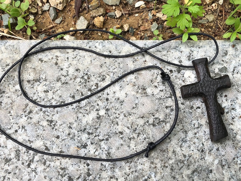 Puede incluir: Un colgante de cruz de madera negra en un collar de cord&oacute;n negro. La cruz tiene una superficie rugosa y texturizada.