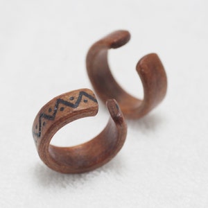 May include: Two wooden rings with a geometric pattern carved into one of the rings. The rings are brown and have a natural wood finish.
