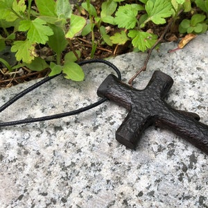 Puede incluir: Un colgante de cruz de madera marr&oacute;n oscuro en un collar de cord&oacute;n negro. La cruz tiene una superficie rugosa y texturizada.