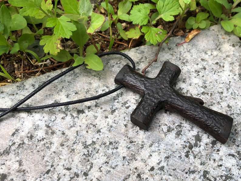 Puede incluir: Un colgante de cruz de madera marr&oacute;n oscuro en un collar de cord&oacute;n negro. La cruz tiene una superficie rugosa y texturizada.