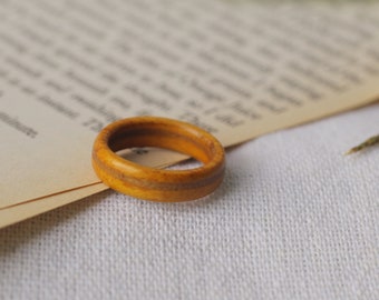 May include: A handcrafted wooden ring with a warm, golden-brown hue. The ring features a simple, classic design with a visible grain pattern. The ring is resting on a vintage paper background.
