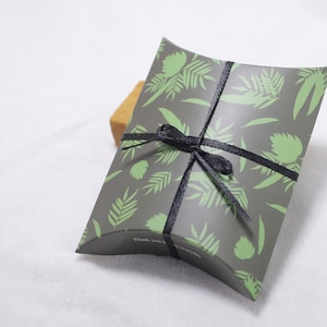Puede incluir: Una caja de almohada gris con un patr&oacute;n de hojas verdes y una cinta negra atada alrededor. La caja tiene el texto "Gracias" impreso en ella.