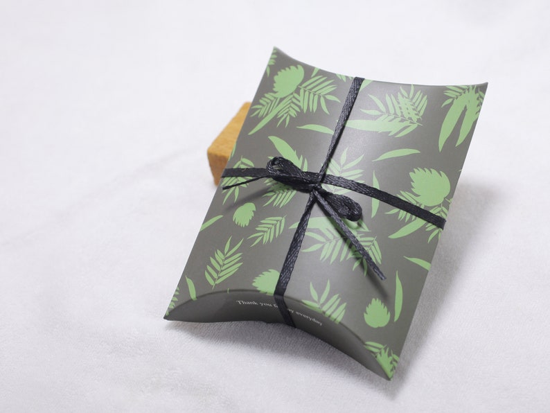 Puede incluir: Una caja de almohada gris con un patr&oacute;n de hojas verdes y una cinta negra atada alrededor. La caja tiene el texto "Gracias" impreso en ella.