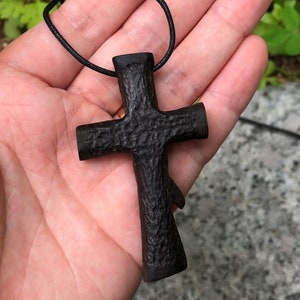 Puede incluir: Un colgante de cruz de madera negra en un cord&oacute;n negro. La cruz est&aacute; tallada con una superficie rugosa y texturizada.