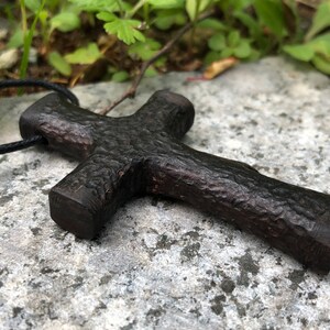 Puede incluir: Un colgante de cruz de madera marr&oacute;n oscuro con una superficie rugosa y texturizada. La cruz est&aacute; unida a un cord&oacute;n negro.