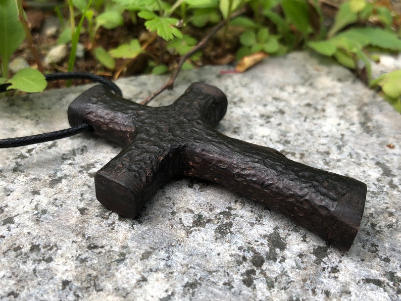 Puede incluir: Un colgante de cruz de madera marr&oacute;n oscuro con una superficie rugosa y texturizada. La cruz est&aacute; unida a un cord&oacute;n negro.