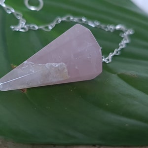 Könnte beinhalten: Ein rosa Rosenquarz-Kristallpendel an einer silbernen Kette. Das Pendel hängt an einem grünen Blatt.