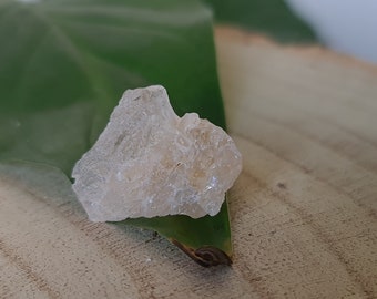 May include: A small, clear, crystalline rock with a rough, irregular surface. The rock is resting on a green leaf.