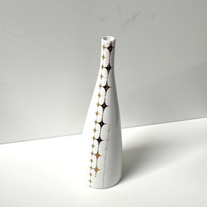 May include: A white ceramic vase with a gold and black geometric pattern. The vase has a narrow neck and a wide base.