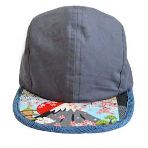 Peut inclure: Une casquette grise à 5 panneaux avec une visière en denim bleu. La visière présente un imprimé coloré d'inspiration japonaise avec le mont Fuji, des fleurs de cerisier et un soleil rouge.