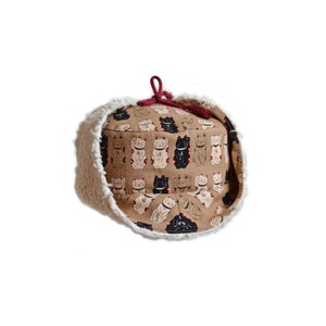 Könnte beinhalten: Ein brauner und weißer Bucket-Hut mit einem weißen Kunstfellbesatz. Der Hut ist mit einem Muster aus schwarzen und weißen Glückskatzenfiguren verziert.